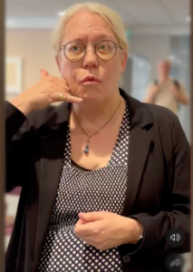 Gunilla Karlsson står i en ljus lokal och tittar mot kameran. Hon tecknar telefon.